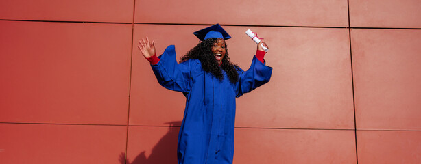Joyful african american woman in graduation cap and gown holding a diploma and cheering