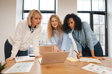 Diverse businesswomen collaborating on project using laptop in modern office discussing