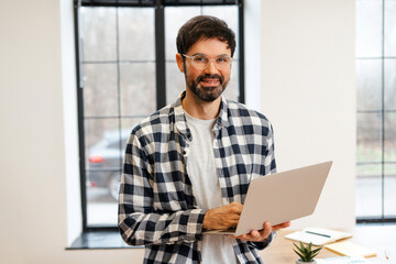 Fototapeta premium Smiling bearded man wearing stylish eyeglasses working remotely using laptop in home office