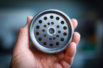 Hand holding a metal component with multiple circular holes arranged in concentric patterns against a blurred background for industrial or mechanical use concepts