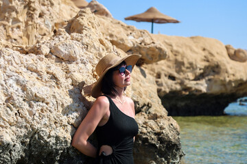 A woman in black swimsuit and hat stands in the water on a beach next to rocks.