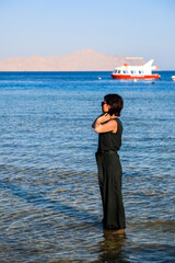 A grown woman in casual clothes stands knee deep in ocean water.