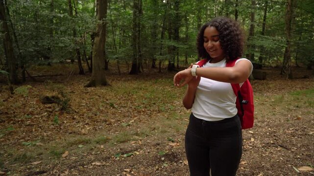 Teenage mixed race African American girl young woman hiking with a red backpack and wearing a smart watch in forest woodland 
