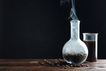 Glass flask with coffee, beans and beaker in smoke on wooden table against black background, closeup. Space for text