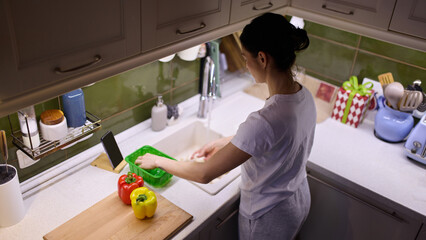 Woman preparing vegetables in modern home kitchen with smartphone on counter. Concept of everyday cooking routine, smart home lifestyle, digital assistance in daily tasks, domestic technology usage.