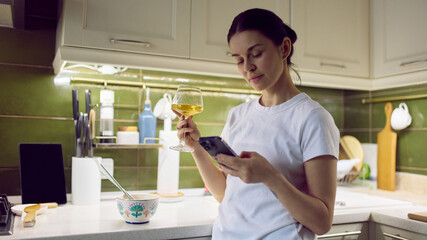 Woman holding glass of wine and checking smartphone in cozy home kitchen. Concept of lifestyle advertising, digital service promotion, home consumption ads and relaxed evening marketing content.