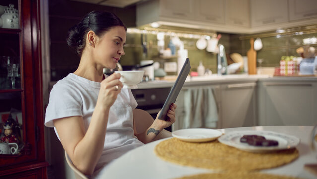 Woman using tablet while drinking coffee at kitchen table at home. Concept of lifestyle advertising, digital content promotion, online services ads and relaxed home media consumption. - Powered by Adobe