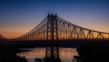 Majestic steel suspension bridge at sunset with reflective river and warm sky