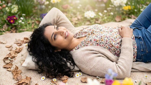 A South Asian woman lying on a picnic blanket in a blooming spring park, looking up at the sky with a peaceful smile.