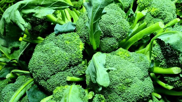 A close-up of fresh broccoli heads piled at a vegetable market, showing dense green florets, thick stems, and crisp leaves that indicate recent harvest and freshness.