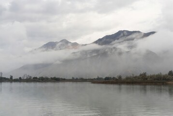 Serene mountain lake landscape with peak partially hidden by thick white fog and low clouds. Moody overcast wilderness scene with calm water reflecting a misty mountain range in grey tones.