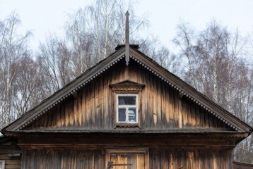 A close-up of a wooden house with a gable decorated with carved cornices and a small window. Vintage rustic architecture of weathered logs against a backdrop of bare birch trees.