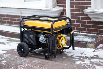 Yellow portable gasoline generator on wheels standing on a snowy sidewalk. Industrial backup power source for home or business during winter blackouts.