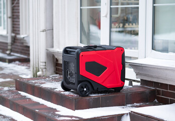 Red portable inverter generator standing on snowy stairs near a building. Backup power source for...