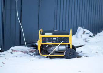Portable yellow power generator standing outdoors in snow near an industrial building, winter emergency electricity supply concept.