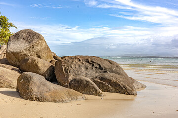 Large weathered rocks on a sandy beach, with calm turquoise water, gentle waves, and a bright blue sky. Nature, travel, sea view, vacation lifestyle imagery.