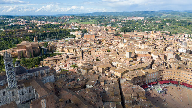Aerial view of the Piazza del Campo's unique shell shape contrasting with the Duomo's dark dome, a tapestry of terracotta roofs unfolds beneath a bright sky, Siena, Tuscany, Italy.