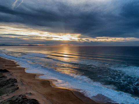Aerial view of golden sunlight slicing through heavy clouds over the ocean's foamy waves meeting the sandy beach, Labenne, France.