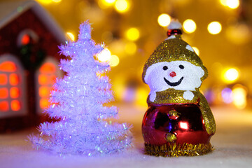A snowman with a white Christmas tree and a small red house on a blurred background with yellow lights