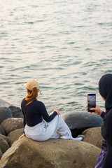a woman takes a picture of her friend posing on the rocks at the beach