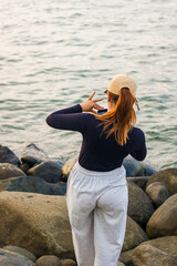 a young woman stands on the rocks on the beach holding her cell phone to take a photo.