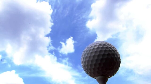 Golf ball on a slender tee rises against a bright blue sky dotted with soft white clouds. Above sky!