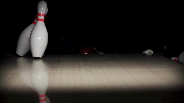 A row of bowling pins stands and leans as red ball rolls near scattered pins, on a glossy floor now