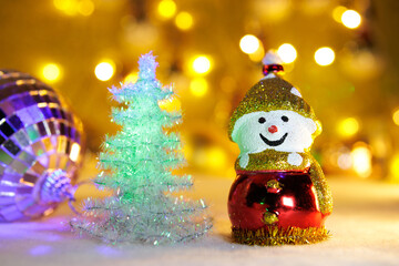 A Christmas tree between a snowman and a Christmas ball on a blurred background with yellow lights