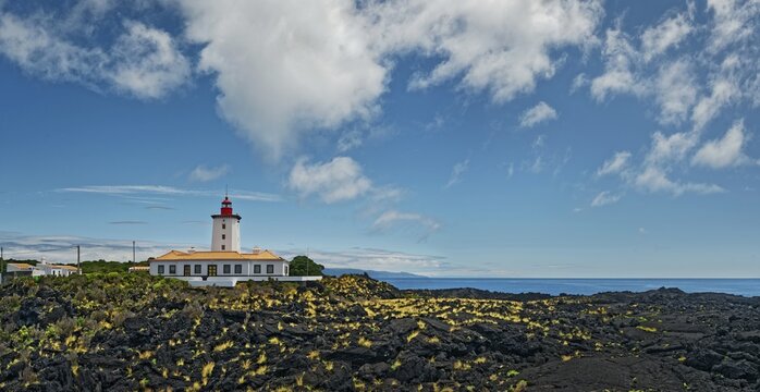 Wide lava flow with the striking lighthouse Farol da Ponta da Iha in front of a blue sky with white clouds, lava rocks coastal path Ponta da Iiha, Manhenha, west coast, Pico, Azores
