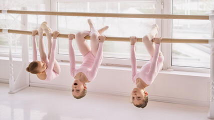 Young ballet students hanging on barre during stretching exercise in studio. Concept of flexibility practice routines, children coordination development, and training session materials.