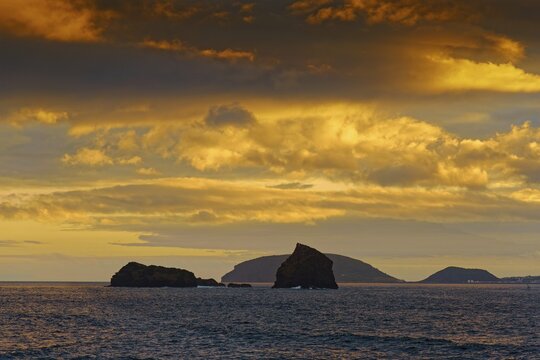 Sea view of the rocks Ilh&eacute;u em P&eacute; and Ilh&eacute;u Deitado under an amber-coloured evening sky with clouds, Madalena, Pico, Azores, Portugal