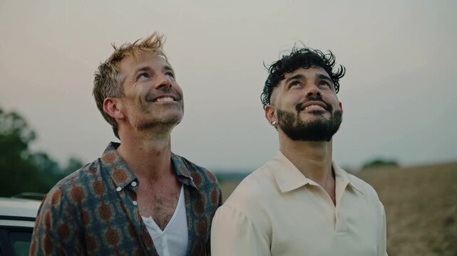 Two joyful men, celebrating LGBTQ+ love, gaze skyward with bright smiles against a soft cloudy backdrop, radiating happiness, warmth, and shared contentment.