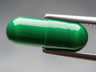 Green capsule held by metal tweezers on gray background  closeup view of medical supplement