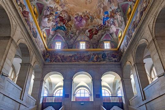 The grand Great Staircase of El Escorial in Spain features massive granite arches leading to a stunning vaulted ceiling covered in vibrant Renaissance frescoes