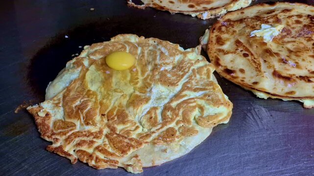 A close-up of a eggroll paratha cooking on a hot griddle with an egg cracked on top, showing set egg whites, a visible yolk, and golden-brown, crisped bread beside another eggroll paratha.