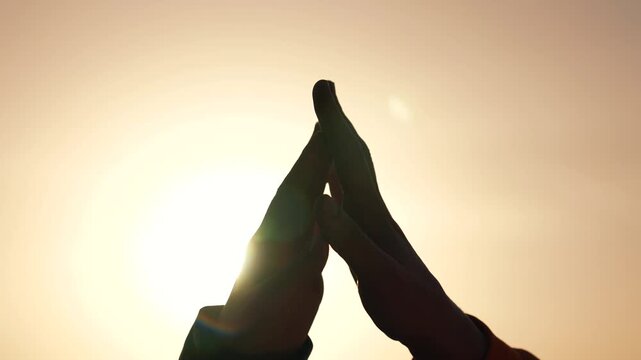 Hands praying in silhouette against sun at sunset, prayer gesture framed by warm sky, spiritual composition conveying unity and faith, hope and meditation emphasis with glow around touching fingers