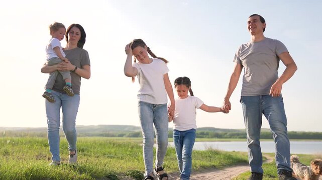 Family walking hand in hand along grassy lakeside path with father mother daughter son child and dog smiling together under clear sunny sky relaxed pace joyful outdoor family walk and playful pet