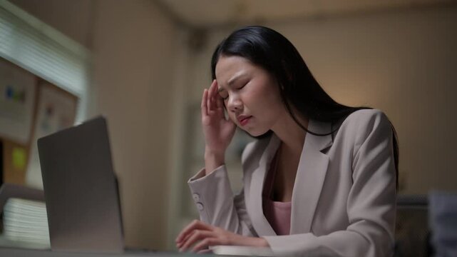 Overworked young asian woman feeling stressed and tired while working on her laptop at the office late at night, suffering from a headache