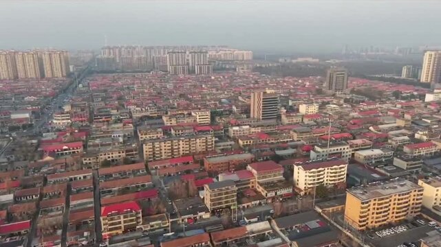 Drone footage showing the residential cityscape of a developing town in central China under hazy weather conditions. The video features dense rows of housing with red roofs and distant high-rise apart