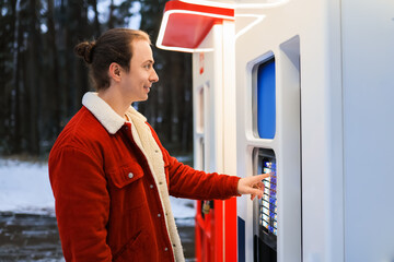 Young man in red winter jacket using outdoor vending machine kiosk in snowy park.
