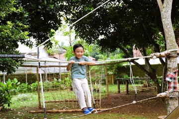 Young child practicing balance and coordination while walking on a rope bridge in a green outdoor playground, promoting mindful movement and sustainable childhood development.