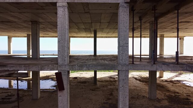 An abandoned building stands partially completed, its raw concrete framing exposed to the elements. The sea glimmers in the distance, creating a striking contrast with the desolate space.