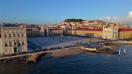 Aerial Lisbon waterfront plaza at sunset, sweeping Tagus river and Praca do Comercio facades bathed in golden light, terracotta rooftops, arcades and cobblestone square with strolling