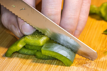 Fresh red, green, and yellow bell peppers are being chopped with a knife on a wooden cutting board. The scene shows healthy cooking, food preparation, and colorful vegetables in a kitchen setting.