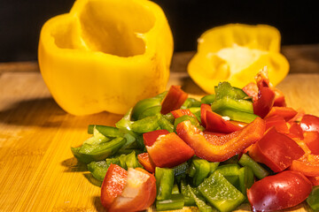 Fresh red, green, and yellow bell peppers are being chopped with a knife on a wooden cutting board. The scene shows healthy cooking, food preparation, and colorful vegetables in a kitchen setting.