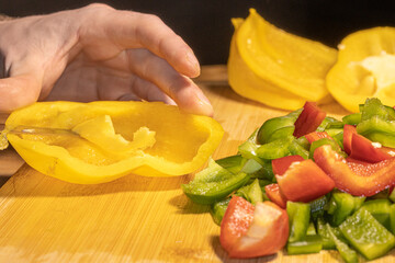 Fresh red, green, and yellow bell peppers are being chopped with a knife on a wooden cutting board. The scene shows healthy cooking, food preparation, and colorful vegetables in a kitchen setting.