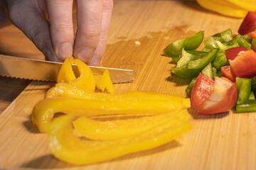 Fresh red, green, and yellow bell peppers are being chopped with a knife on a wooden cutting board. The scene shows healthy cooking, food preparation, and colorful vegetables in a kitchen setting.