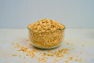 Abundant golden rolled oats overflowing from a clear glass bowl sitting on a textured light surface ready for healthy breakfast preparation