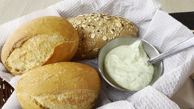 Fresh bread rolls in a basket with white paper, including wheat and multigrain buns, served with a bowl of creamy herb butter or dip. Concept of breakfast, bakery, homemade food.