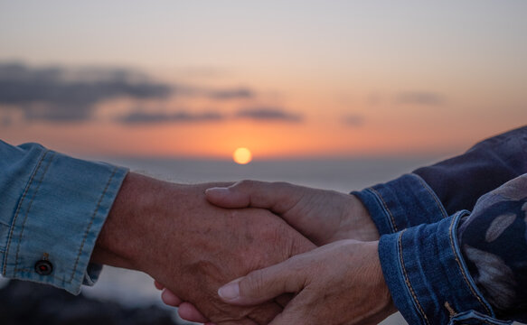 Two old female hands holding a man's hand, concept of protection, love, support, protection and help. In the background the sun sets into the sea